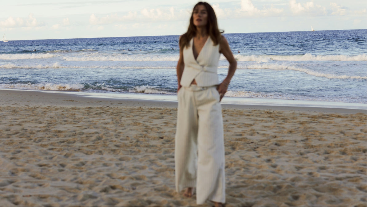 A woman stands on a sandy beach wearing a white sleeveless wrap-style top and matching wide-leg trousers, with the ocean and sky behind her.